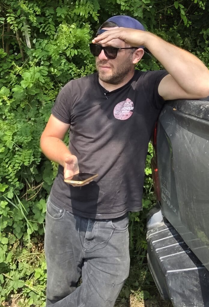 David Large, le vigneron du domaine éponyme, se tient debout, appuyé sur l'arrière d'un pick-up foncé. Il porte un t-shirt noir, un jean noir, une casquette de baseball bleue retournée et des lunettes de soleil noires. Il a une barbe. Il protège ses yeux de la main droite. Dans sa main gauche, il tient un téléphone portable avec un étui doré. Derrière lui, il y a des buissons et des arbres. La lumière du soleil est vive.
